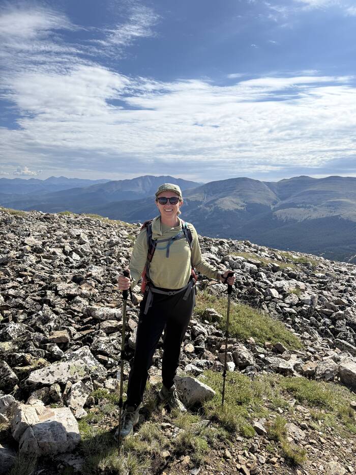 Nikki Bisel smiling on a rocky ridge above treeline, holding trekking poles, with mountain ranges and a wide blue sky behind her.